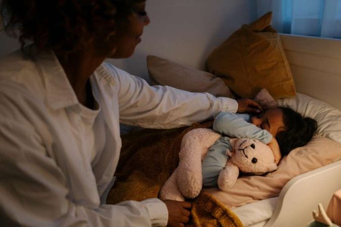 Une mère se penche avec tendresse pour caresser les cheveux de son jeune enfant qui dort paisiblement dans un lit avec une peluche.