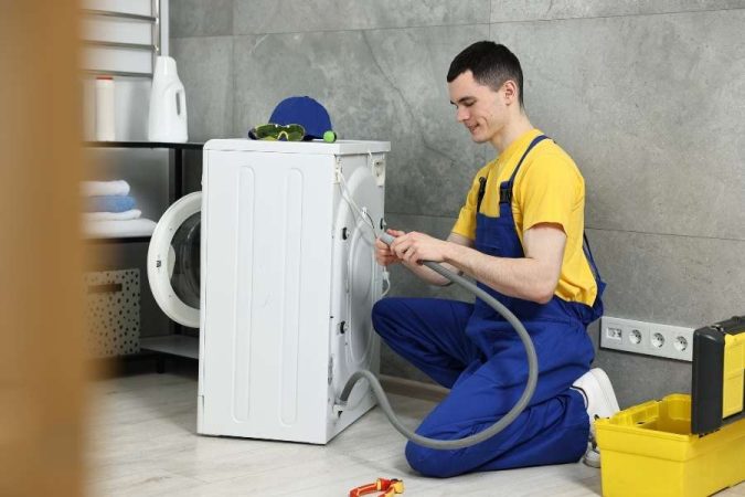 Un technicien en combinaison bleue installe ou vérifie le tuyau d'évacuation de la machine à laver dans une buanderie.