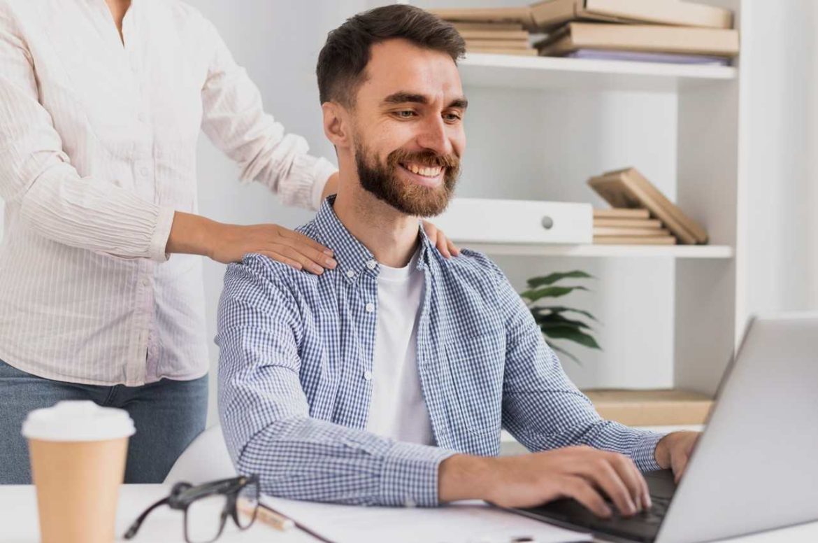 Un homme souriant travaillant sur son ordinateur pendant qu'une femme lui masse les épaules au bureau lors d'une séance de massage amma assis pause détente rapide.
