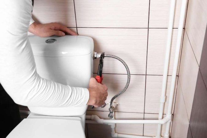 Un plombier répare une fuite de chasse d'eau dans des toilettes carrelées.